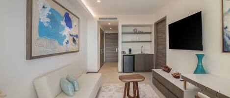 Una sala de estar moderna con sofá, una mesa de centro, televisor y una zona de cocina con refrigerador y estanterías.