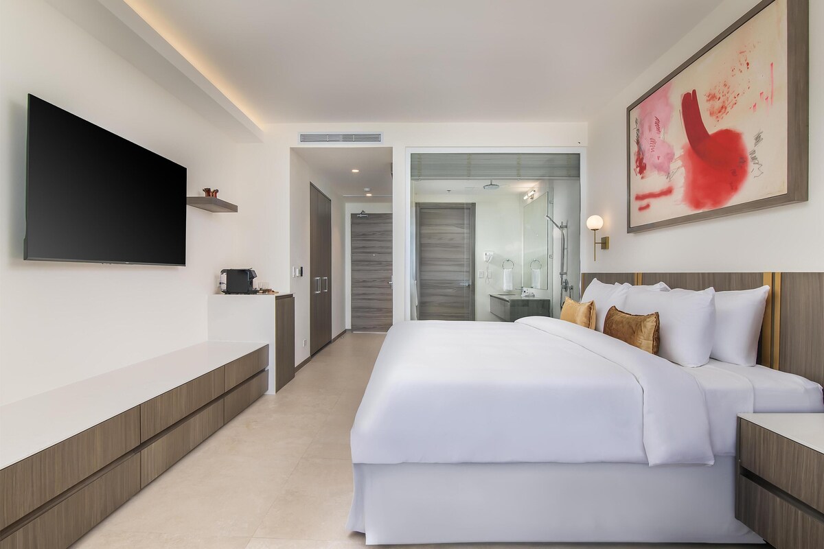 Una habitación de hotel moderna con una cama grande, un televisor de pantalla plana, una mesita de noche y un baño visible a través de una puerta abierta.