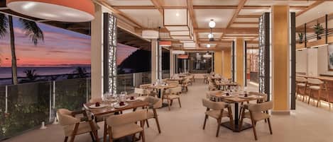 Un comedor amplio con varias mesas y sillas, vistas al atardecer y un balcón con barandillas de vidrio.