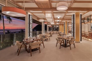 Un comedor amplio con varias mesas y sillas, vistas al atardecer y un balcón con barandillas de vidrio.