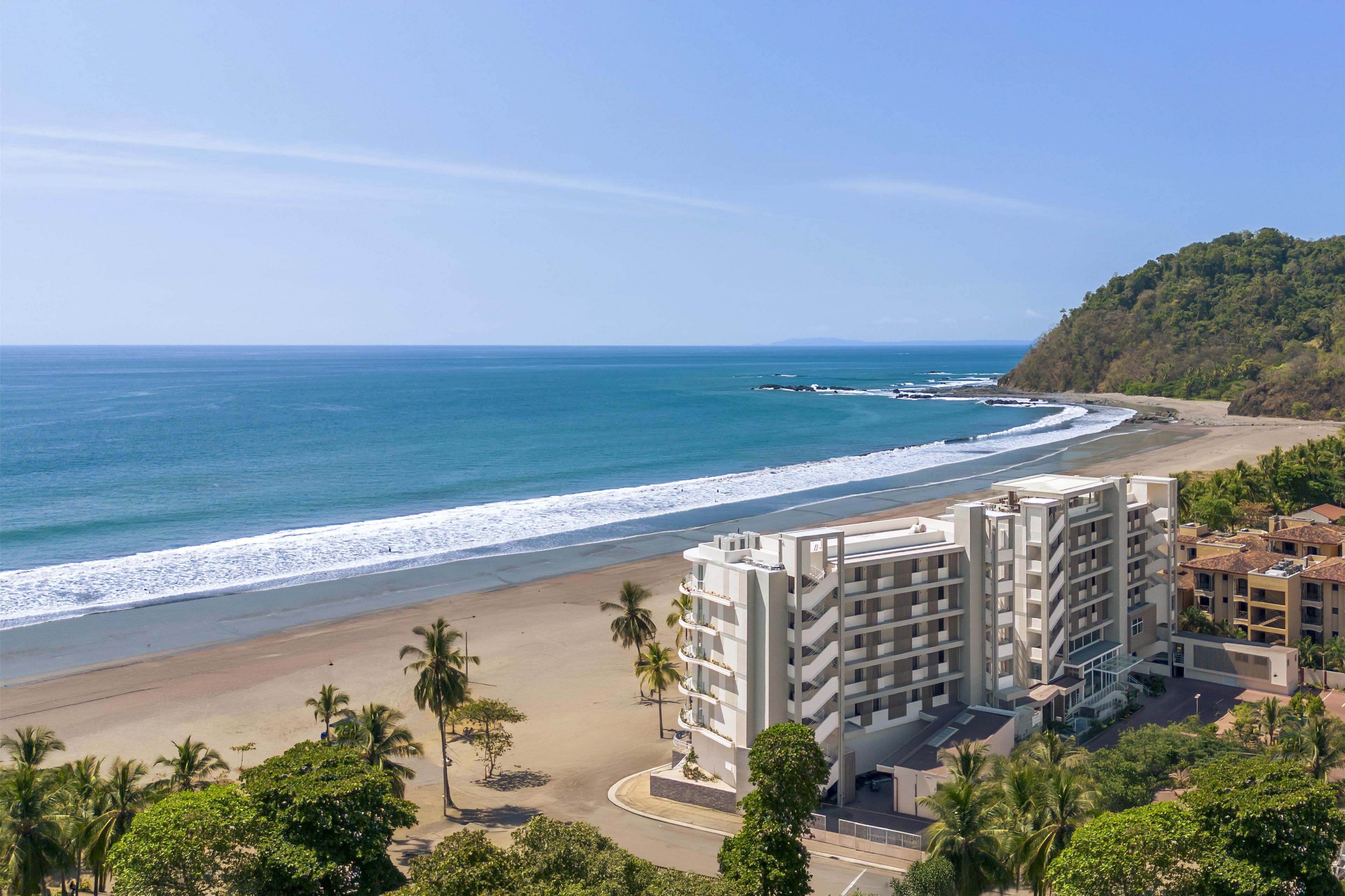 Un hôtel situé sur la côte, composé de plusieurs bâtiments, avec une plage et une colline boisée.