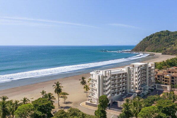 Un hôtel situé sur la côte, composé de plusieurs bâtiments, avec une plage et une colline boisée.
