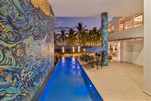A pool with a blue water feature, palm trees, and a modern building with large windows. - Clarita's Beach Bar & Resort, WorldHotels Elite (Jacó)