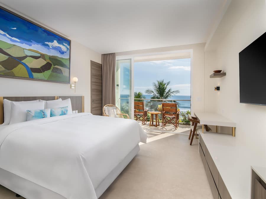 A hotel room with a large bed, a painting, a TV, and a balcony with chairs.
