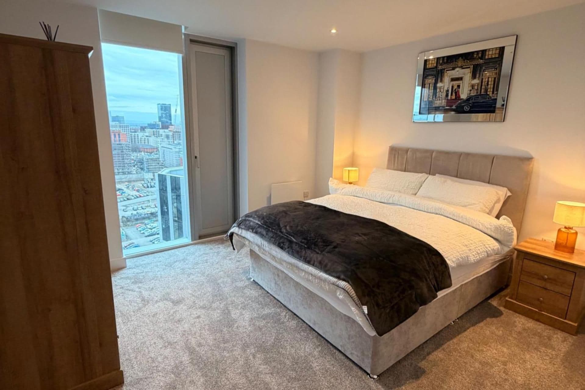 Une chambre avec un grand lit, une armoire en bois, une vue sur la ville à travers une porte coulissante et un tableau encadré au mur.