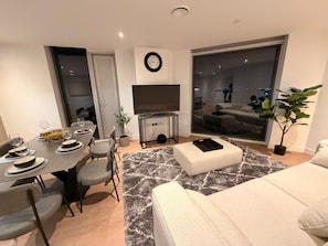 A modern living room with a dining area, a flat-screen TV mounted on the wall, and a large window offering a view of the cityscape. - Modern City Apartment in Manchester (Manchester)