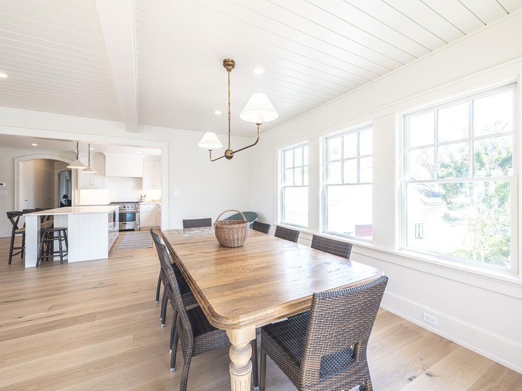 A spacious dining area with a wooden table and wicker chairs, a kitchen in the background, and large windows providing natural light.