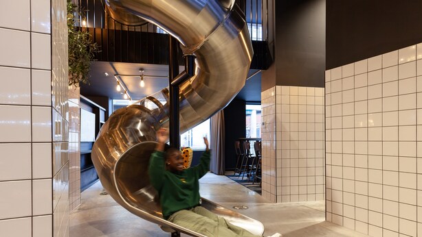 Een persoon glijdt een spiraalvormige metalen glijbaan af in een moderne, overdekte ruimte met betegelde muren en een trap op de achtergrond.