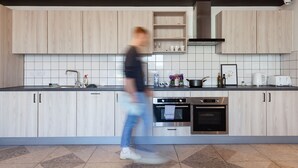 Een moderne keuken met lichte houten kastjes, een betegelde achterwand, roestvrijstalen apparatuur, en een persoon die erdoorheen loopt.