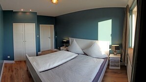 Een slaapkamer met een groot bed, twee witte deuren, een nachtkastje met een lamp en een raam met gordijnen.