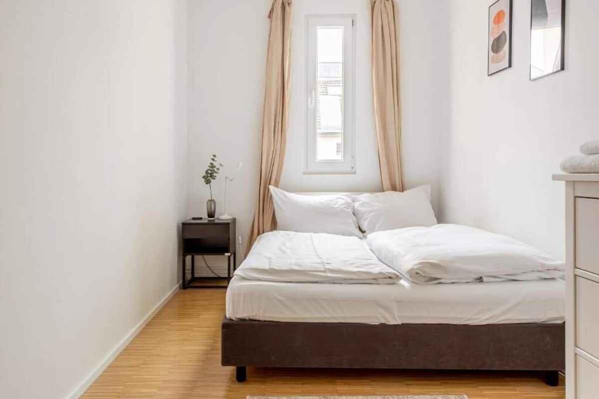 Ein Schlafzimmer mit einem Bett, einem Nachttisch, einem Fenster mit Vorhängen und einer kleinen Pflanze.