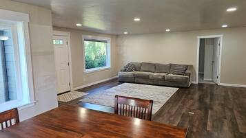 A spacious living room with a large grey sofa, a wooden dining table, and a view of greenery through a window.