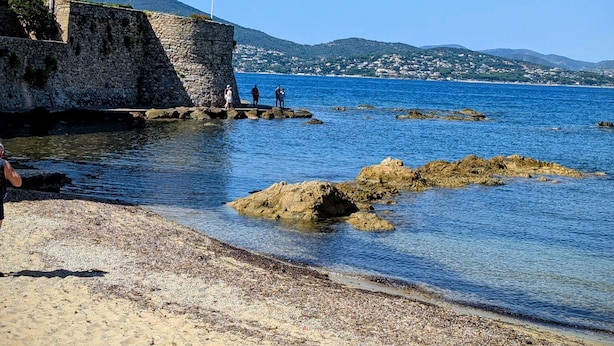 Uma praia costeira com águas azuis cristalinas, afloramentos rochosos e um muro de pedra.