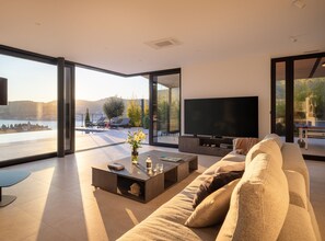 Ein modernes Wohnzimmer mit großem Fernseher, bequemen Sitzmöglichkeiten und Blick auf einen See und Hügel.