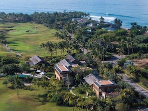 An aerial view of a coastal resort with a golf course, beachfront villas, and lush greenery.