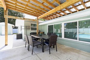 En udendørs terrasse med et træpergola, et spisebord med stole og udsigt til et hus med grøn facade og vinduer.