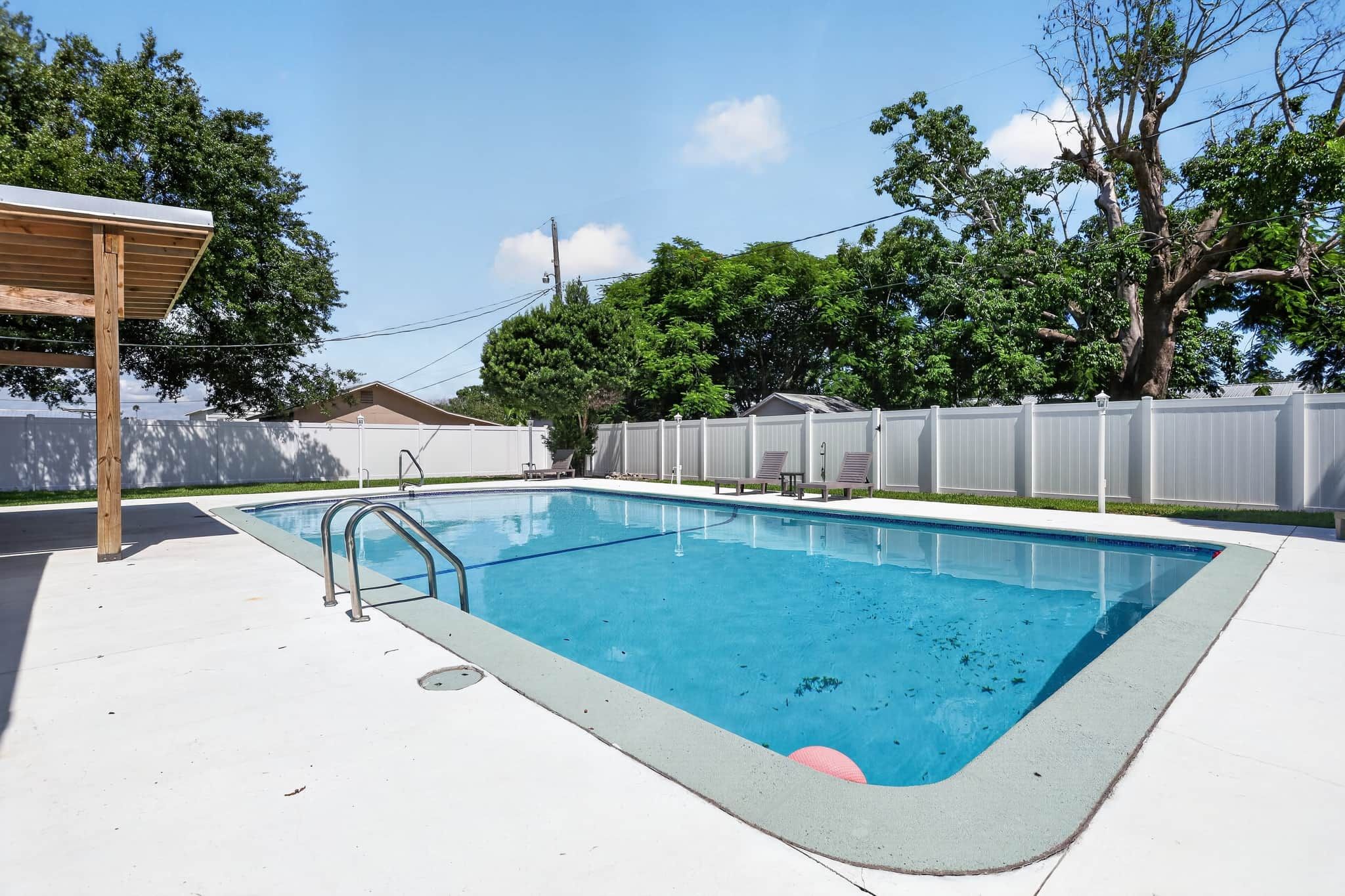 Ein rechteckiger Swimmingpool mit Metallleiter, umgeben von einem weißen Zaun, unter blauem Himmel.