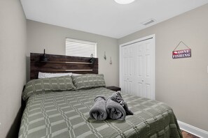 Un dormitorio con cama, mesita de noche, ventana con persianas, puerta y un cartel en la pared.