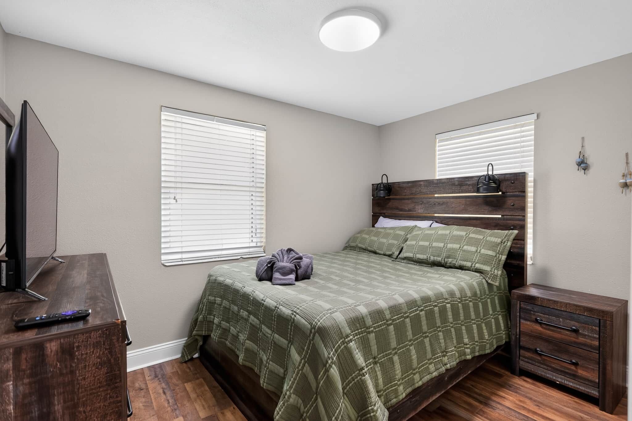 Un dormitorio con cama, mesita de noche, cómoda, televisor y dos ventanas con persianas.