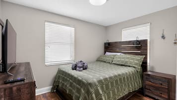 Un dormitorio con cama, mesita de noche, cómoda, televisor y dos ventanas con persianas.