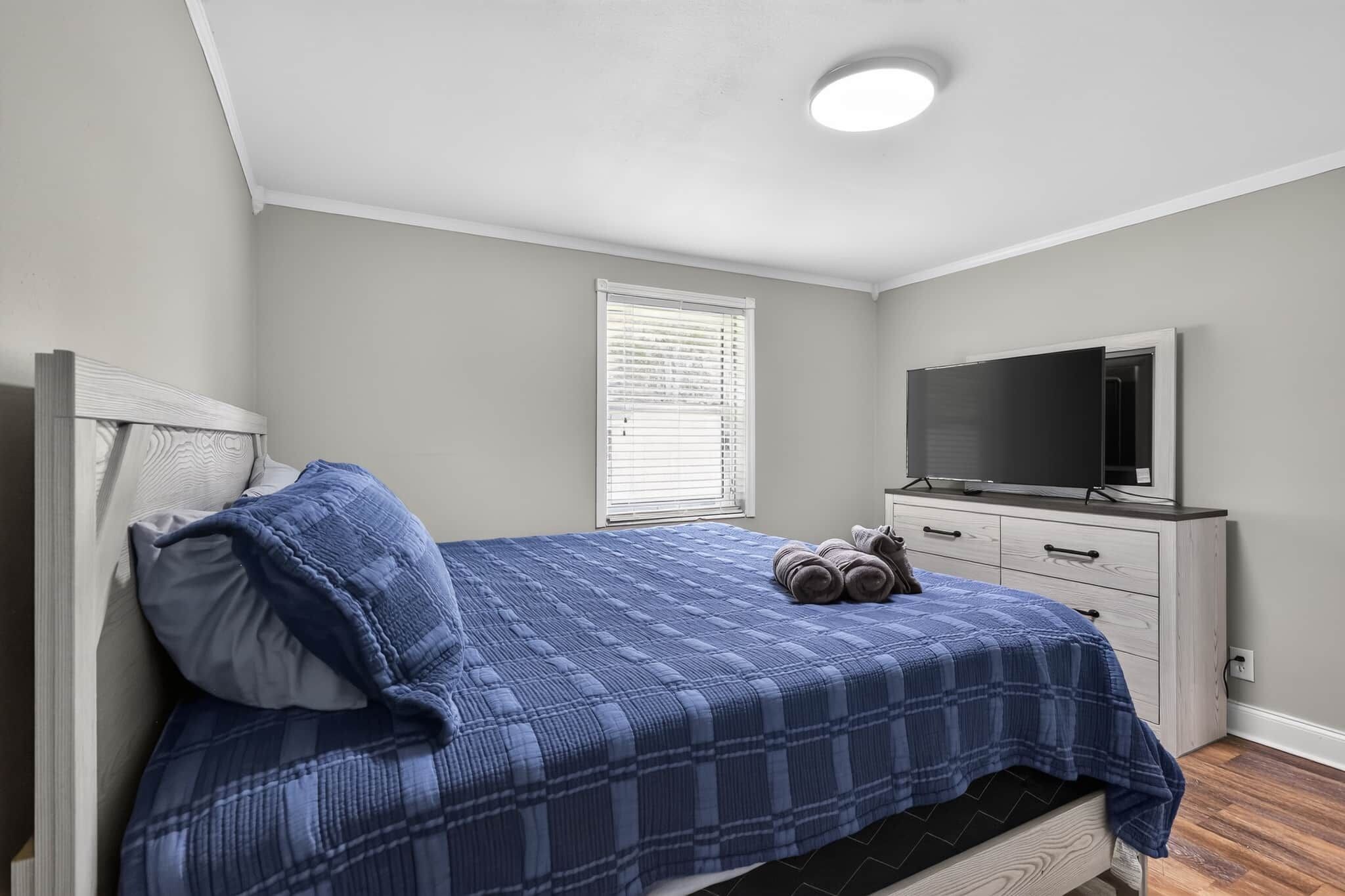 Un dormitorio con cama, televisor, ventana con persianas y cómoda.
