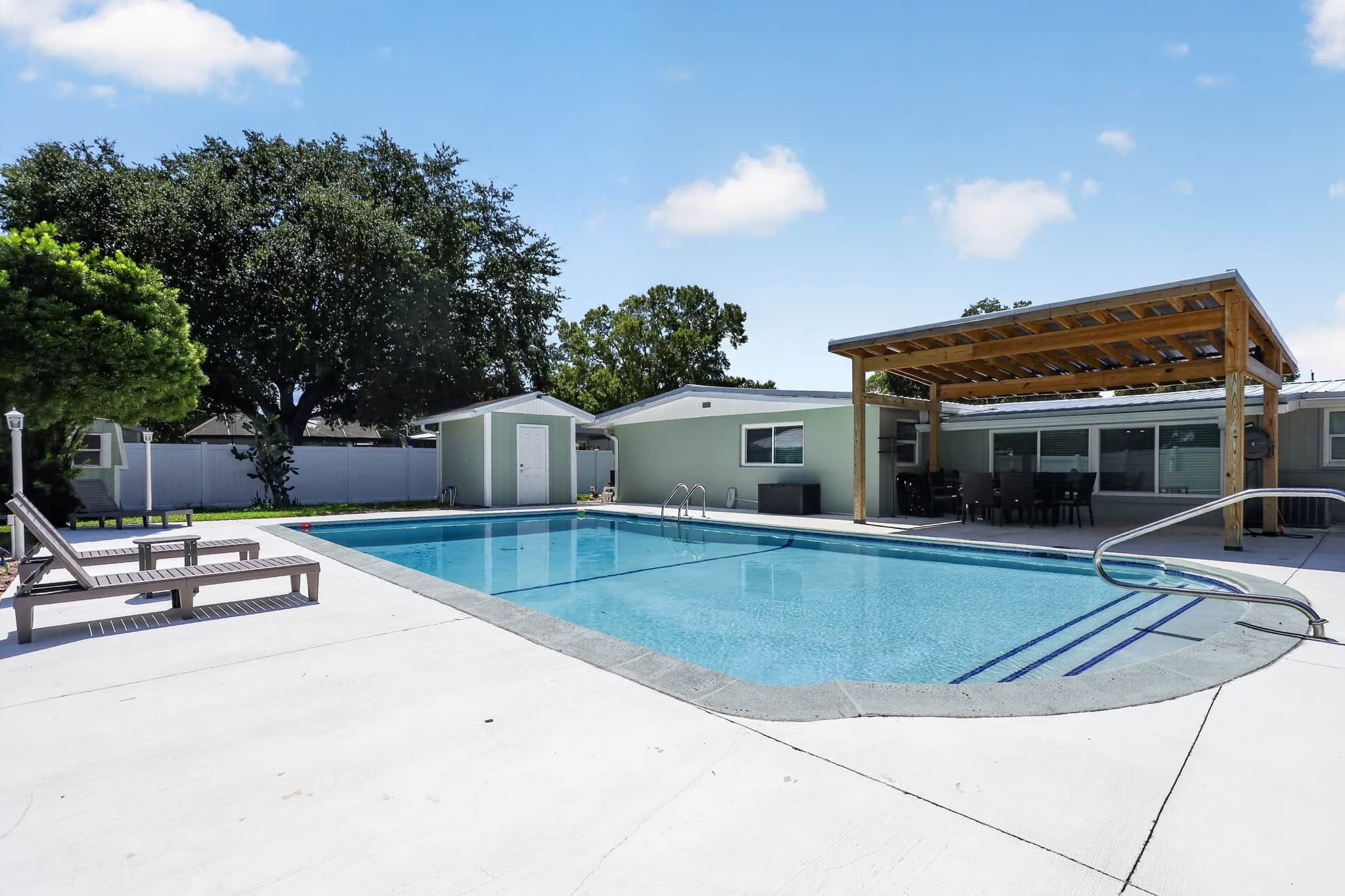 A swimming pool with a wooden deck, lounge chairs, and a covered patio area.