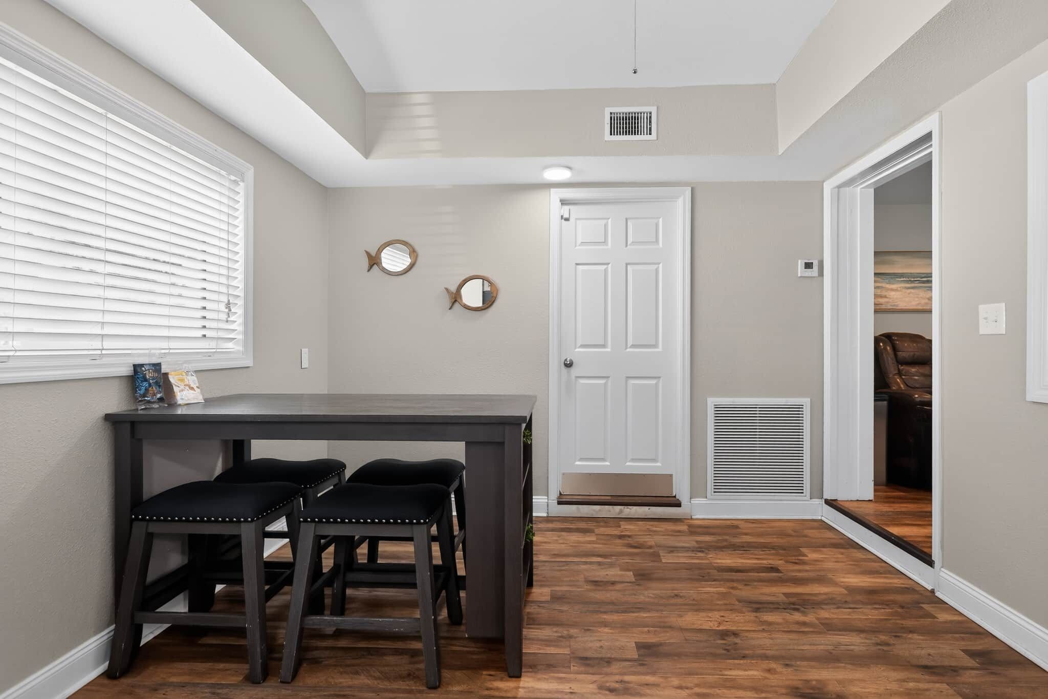 A room with a dining table and chairs, a door, a vent, and a window with blinds.