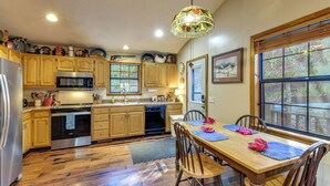 A kitchen with wooden cabinets, stainless steel appliances, a dining table with chairs, and a large window with a view. - Hot Tub & Streamside Bliss! Topton Mtn Haven (Topton)