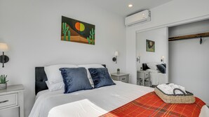 Ein ordentlich eingerichtetes Schlafzimmer mit einem Bett, Kissen, einem Nachttisch mit Lampe, einer kleinen Pflanze und einer wandmontierten Klimaanlage.
