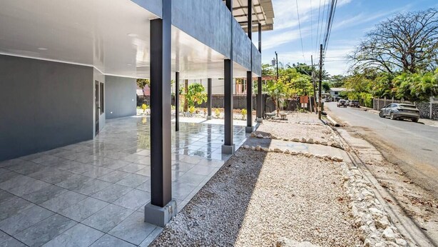 A modern building with a covered outdoor area, a gravel path, and a street with parked cars. - Downtown Apartment by The Beach #5 (Sámara)