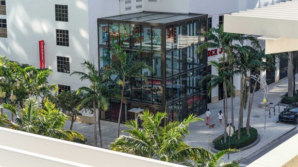 A modern building with a glass facade, palm trees, and a pedestrian walkway. - Midtown Rouge- Condo with City Views, Pool & Gym (Miami)