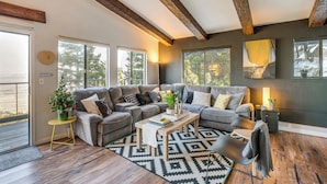 Ein modernes Wohnzimmer mit einer großen Eckcouch, einem Couchtisch und einem gemusterten Teppich. Große Fenster sorgen für natürliches Licht und Blick auf Bäume.