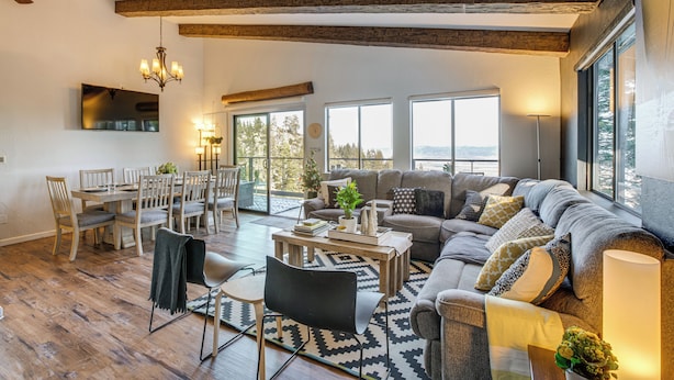 Ein geräumiges Wohnzimmer mit einer großen Couch, einem Esstisch und Stühlen. Ein Kamin, eine Kristallleuchter und große Fenster mit Blick ins Freie sind ebenfalls vorhanden.