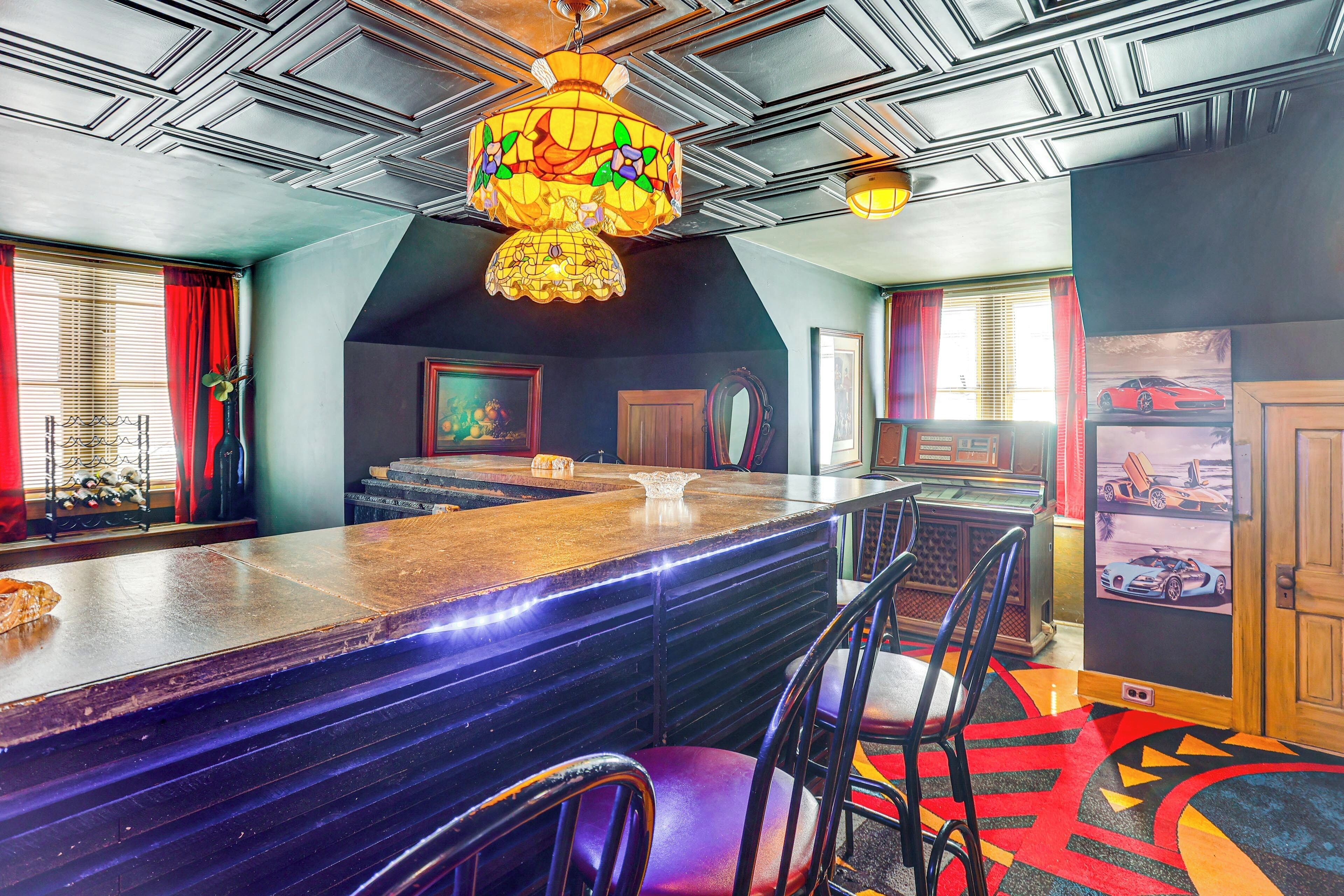 Uma área de bar com um balcão comprido, várias cadeiras, um lustre e um tapete colorido.