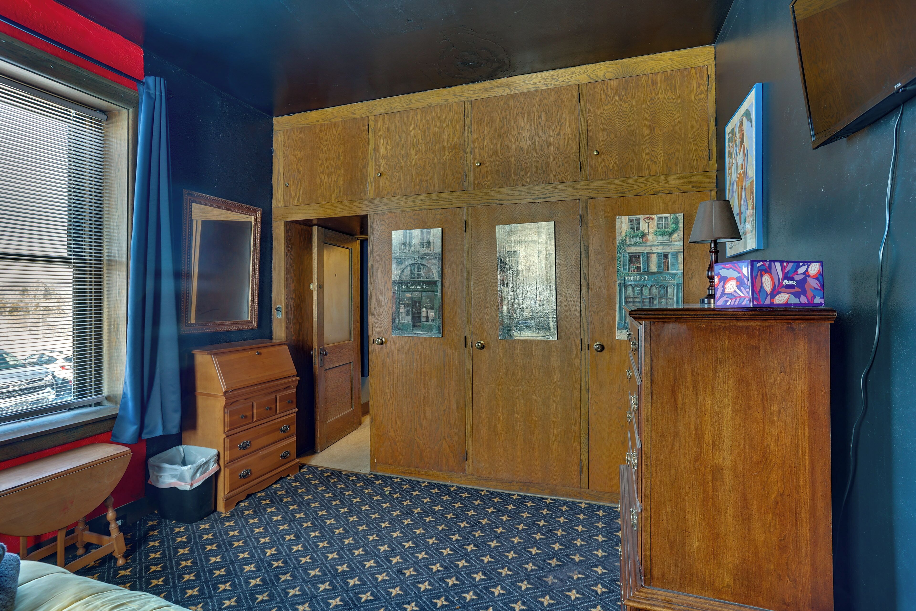 Een kamer met een grote houten garderobe, een bureau met stoel, een kamerbreed tapijt en een raam met jaloezieën.