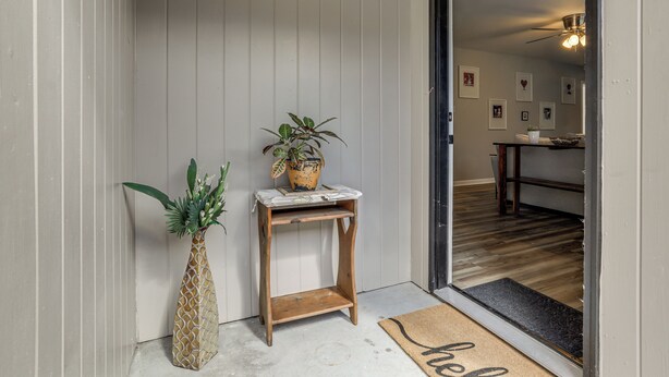 A wooden side table with a potted plant and a vase, a doormat with "hello" on it, and a wooden floor leading to a room with a ceiling fan and framed pictures on the wall. - 1 Mi to Tiger Stadium: Sleek University South Gem! (Baton Rouge)