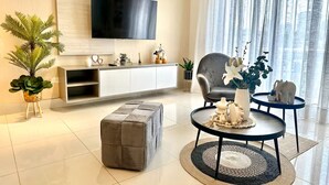 Ein modernes Wohnzimmer mit Wand-Fernseher, einem grauen Sofa, einem runden Couchtisch mit Dekoration und einem weißen Konsolentisch mit Pflanzen und Accessoires.
