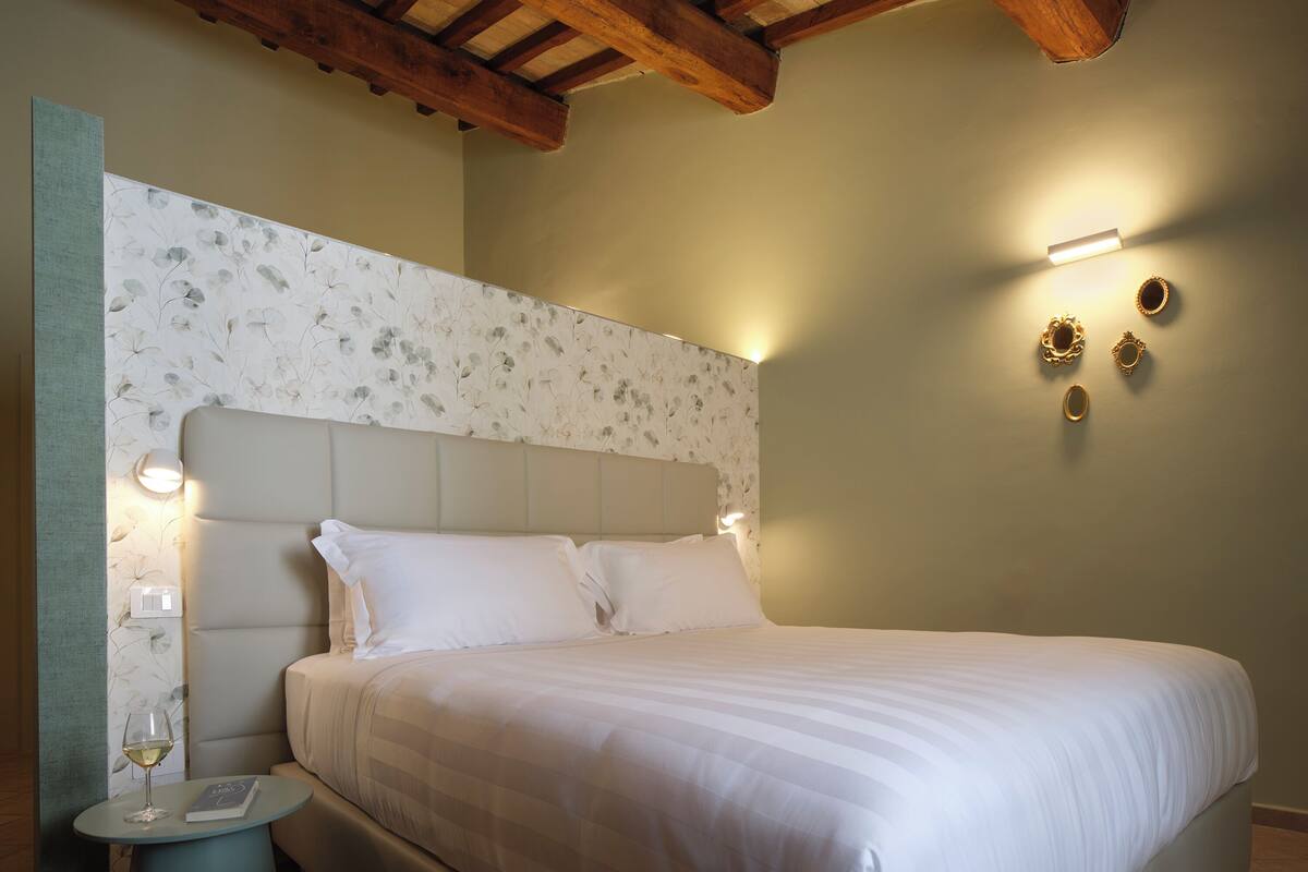 Una cama bien hecha con cabecera, una mesita de noche con una copa de vino y un libro, y luces fijadas en la pared.