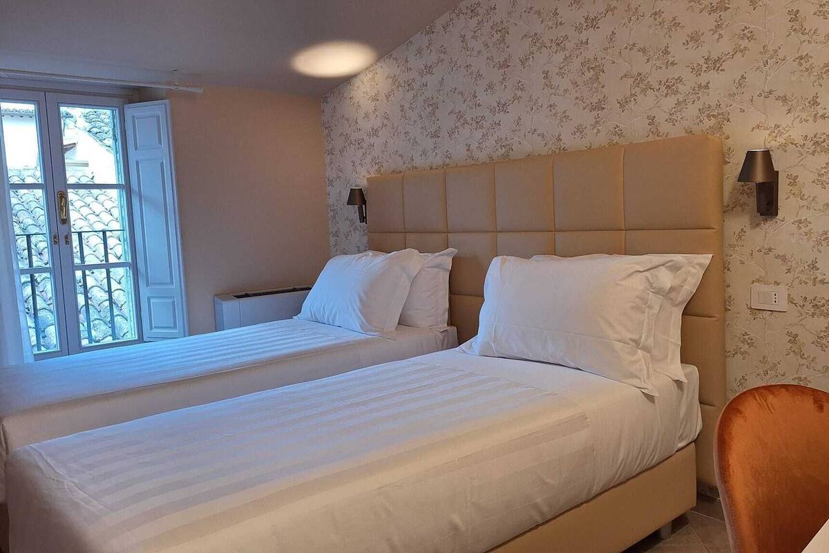 Una cama doble con ropa blanca, cabecera, mesita de noche, lámpara y ventana con vistas.