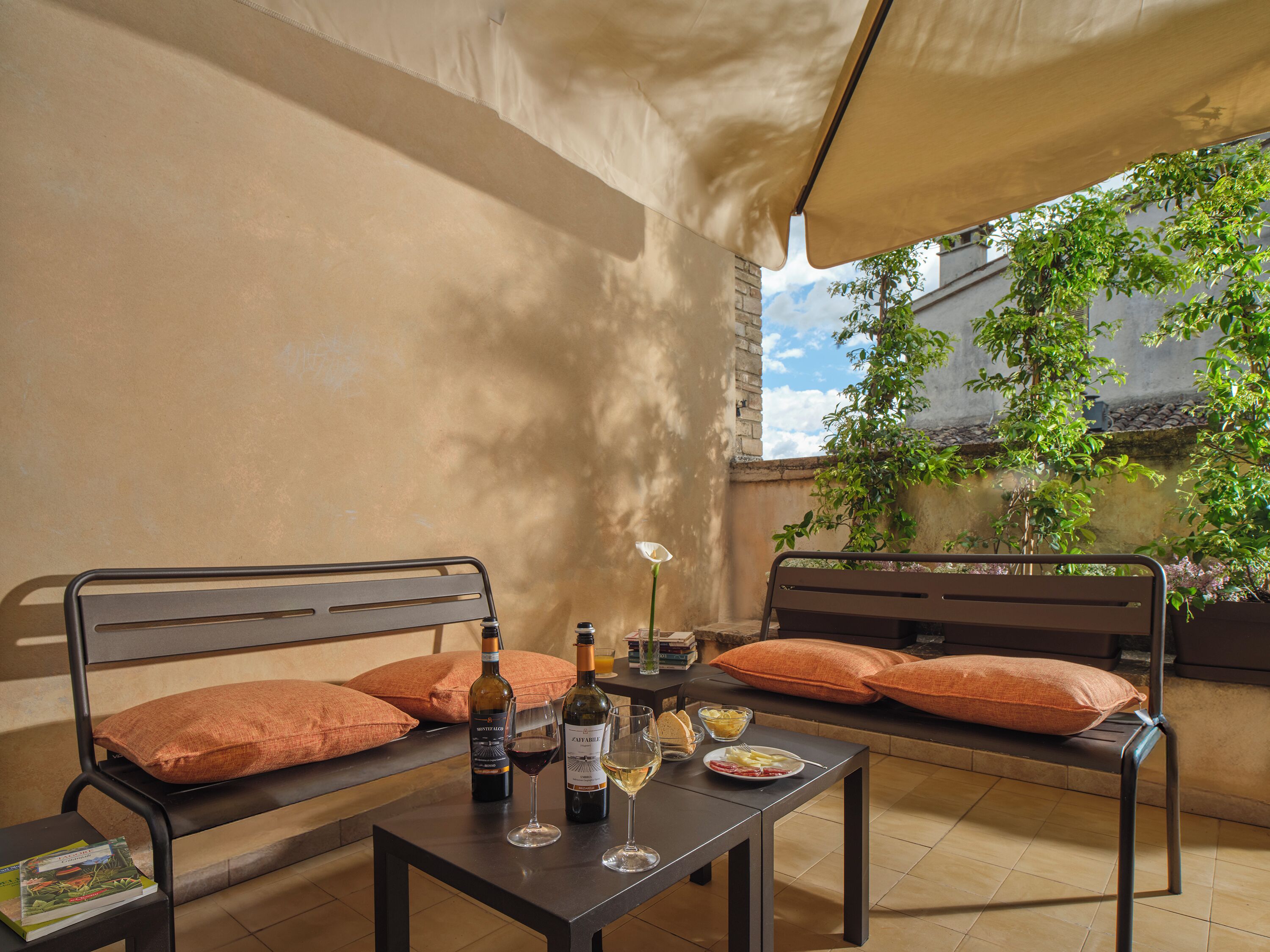 Une terrasse avec deux lits à structure métallique, une table avec des bouteilles de vin et des verres, et une vue sur des bâtiments et des arbres.