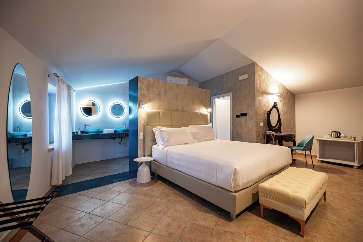 Un dormitorio moderno con una cama grande, una mesita de noche pequeña, una silla y un baño con espejo y lavabo.