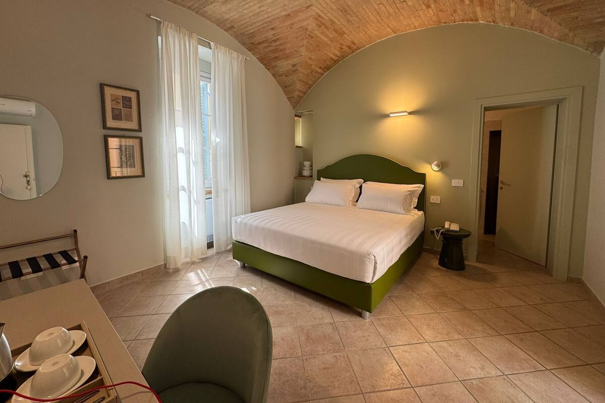 Un dormitorio con una cama verde, un espejo redondo, una silla, una mesa con tazas y una puerta que da a otra habitación.