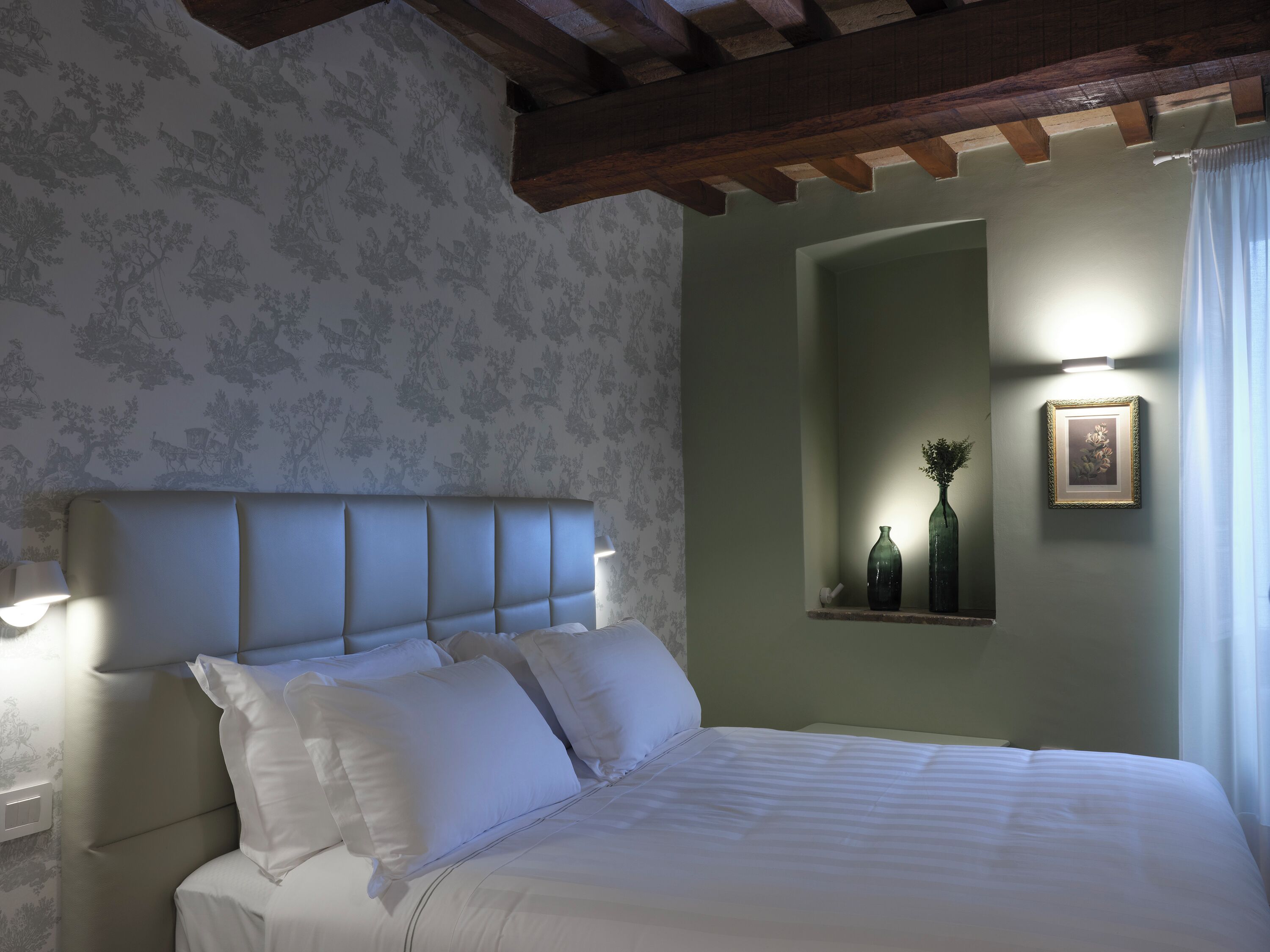 Un dormitorio con cabecera, almohadas, una lámpara de noche, un estante en la pared con botellas y un cuadro enmarcado.