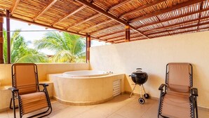 A room with a hot tub, two chairs, a grill, and a view of palm trees.