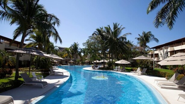 A swimming pool with lounge chairs and umbrellas, surrounded by palm trees and buildings.