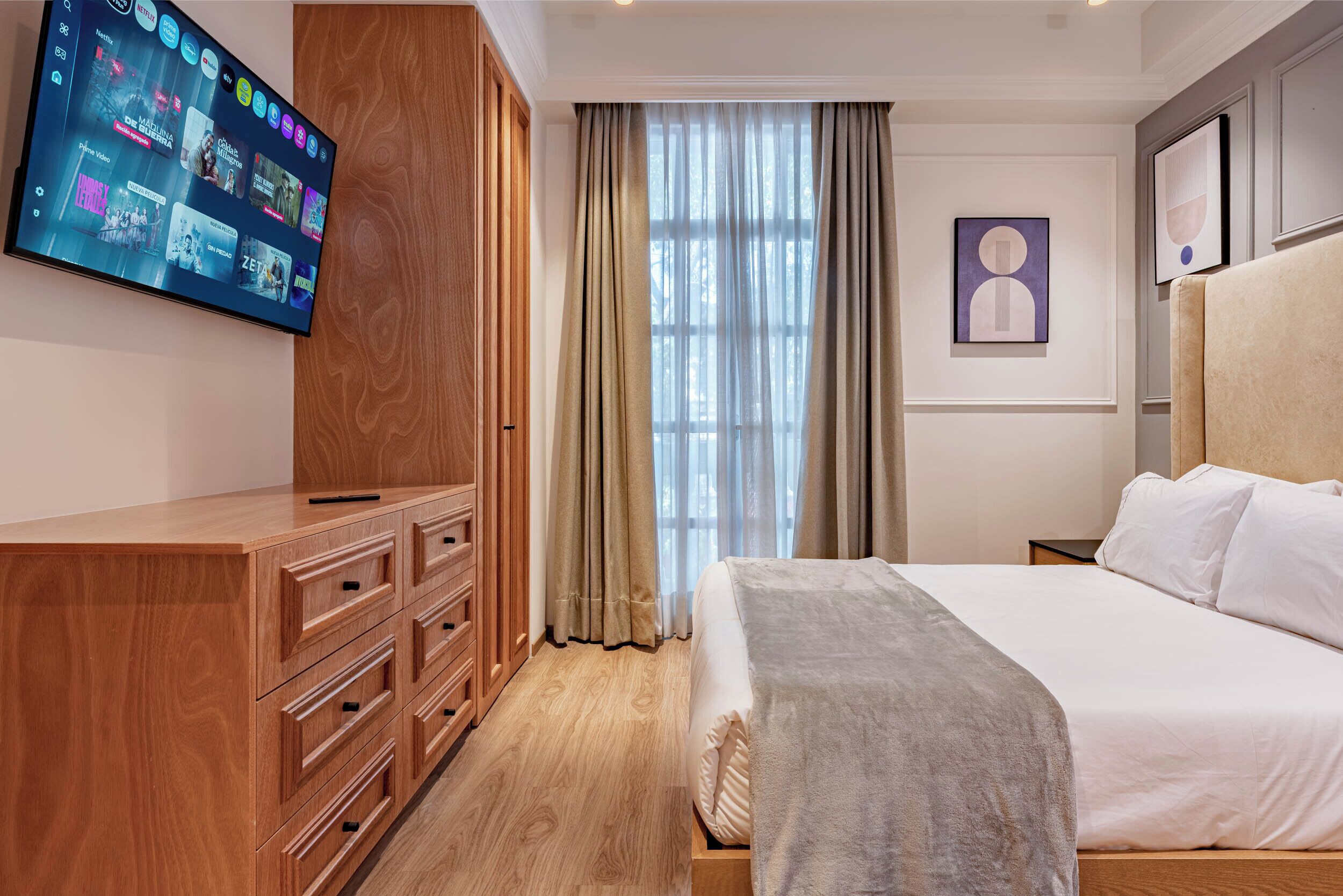 Een hotelkamer met een flatscreen televisie, een houten garderobe, een bed met witte lakens en een groot raam met gordijnen.