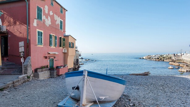 A boat on a pebbled shore, a building with a pink facade, and a calm sea.