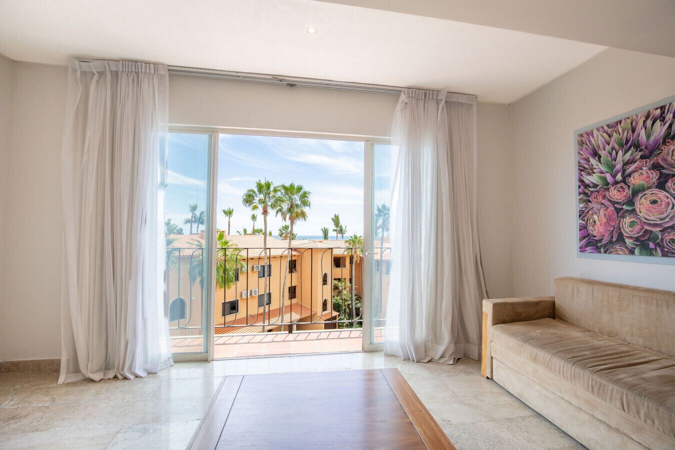 A hotel room with a balcony, a beige sofa, and a painting of flowers.
