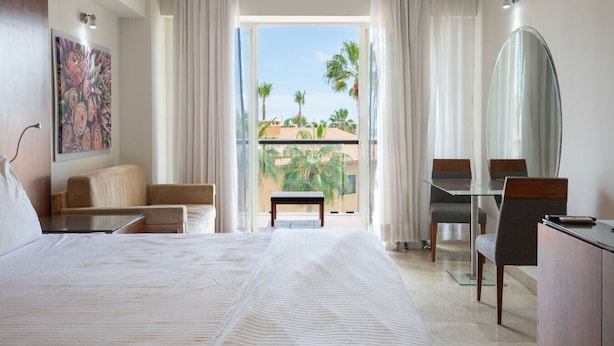 Een hotelkamer met een bed, een bureau, een stoel, een klein tafeltje en een balkon met uitzicht op palmbomen.