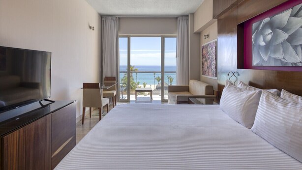 A hotel room with a large bed, a TV, a balcony with a view of the ocean, and a painting on the wall.